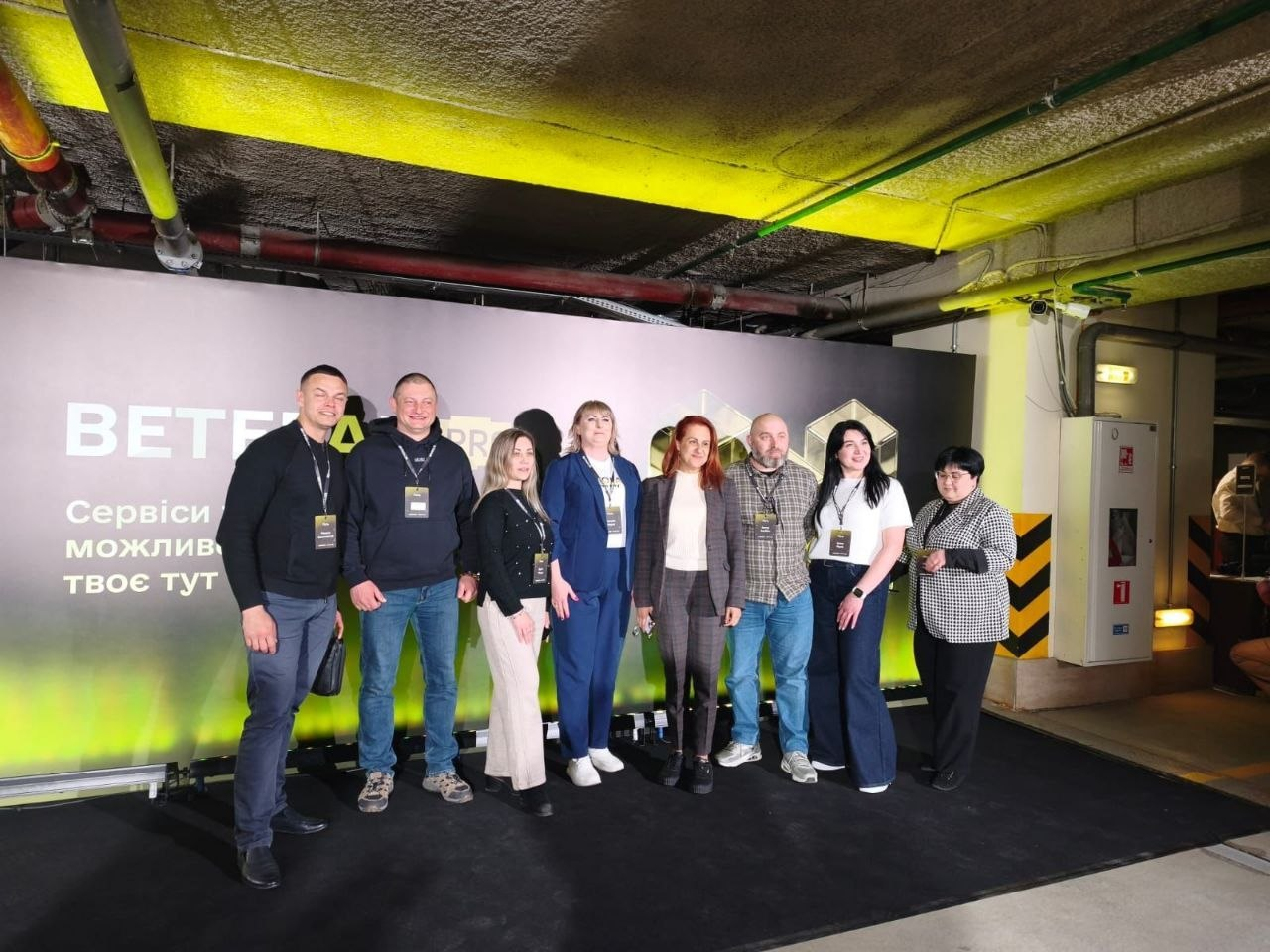 рупа людей стоїть у ряд на фоні банера з написом «BETERA PRO». Усі присутні мають бейджі учасників заходу. Локація нагадує облаштоване підвальне приміщення або паркінг із низькою стелею та відкритими трубами комунікацій.