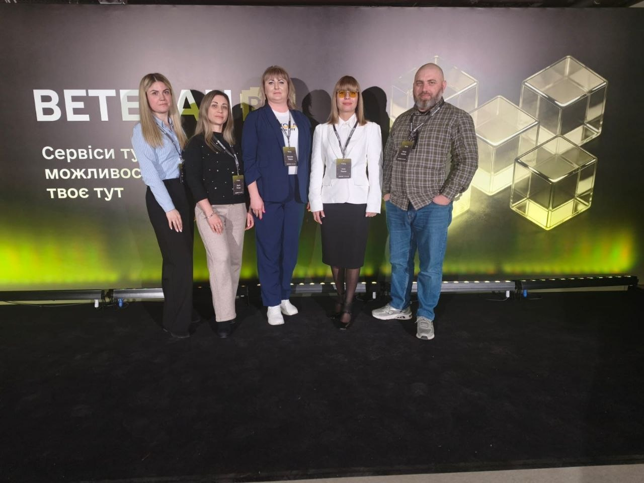 П'ятеро людей стоять на сцені перед темним банером із написом «BETERA PRO» та зображенням скляних кубів. Учасники заходу мають на шиї бейджі. Знизу банер підсвічується яскравою зеленою смугою світла.