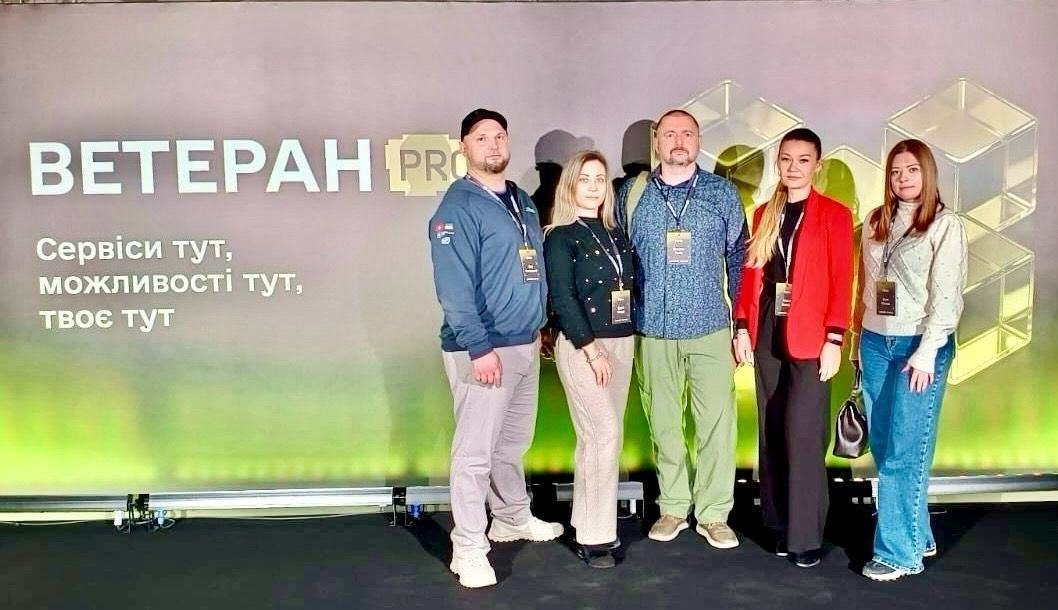 Група з п'яти людей стоїть перед великим банером із написом «ВЕТЕРАН PRO» та гаслом про сервіси й можливості. Усі учасники мають бейджі. Банер оформлений у сірих та жовтих тонах із зображенням скляних кубів.