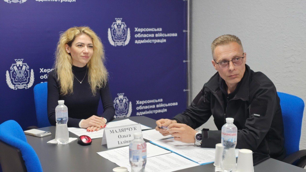 Чоловік та жінка сидять за робочим столом на фоні темно-синього банера з емблемами та написом «Херсонська обласна військова адміністрація». На столі перед ними стоять пляшки води, лежать документи та табличка з ім'ям.