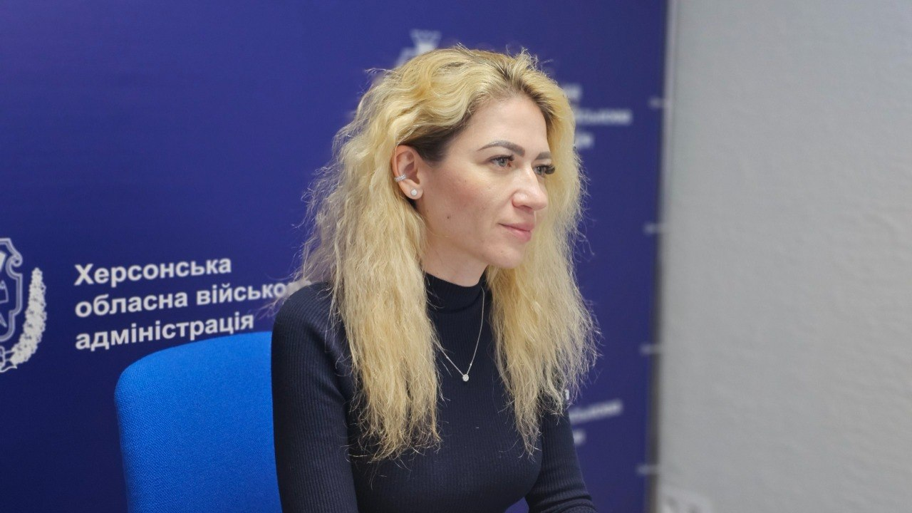 Жінка зі світлим волоссям сидить у профілі на фоні темно-синього банера з написом «Херсонська обласна військова адміністрація». На ній чорна кофта з високим коміром та тонкий ланцюжок із кулоном.