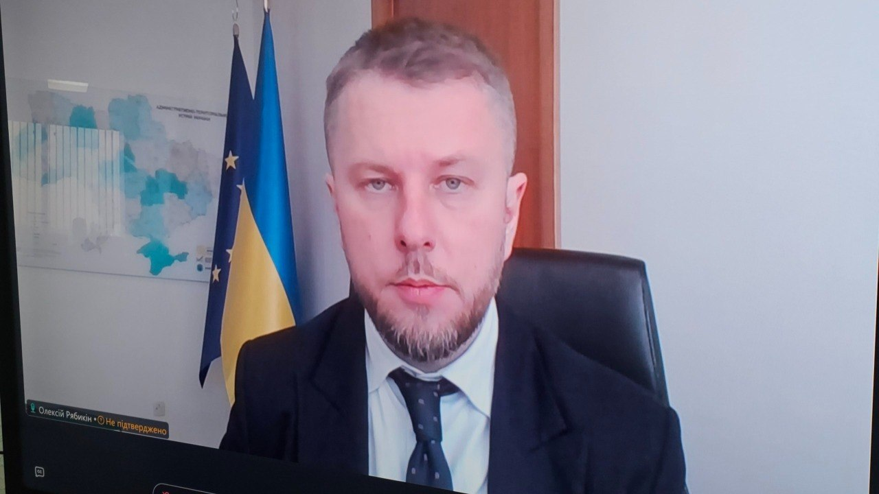 Скриншот відеодзвінка з чоловіком у діловому костюмі та краватці. На задньому плані видно державний прапор України, прапор Європейського Союзу та карту на стіні. У лівому нижньому куті вікна розміщено текстовий підпис.