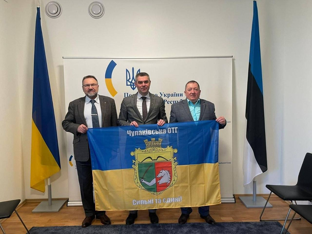 Троє чоловіків у ділових костюмах стоять у приміщенні та тримають прапор Чулаківської ОТГ. На задньому плані розташовані державний прапор України, прапор Естонії та білий стенд із логотипами.