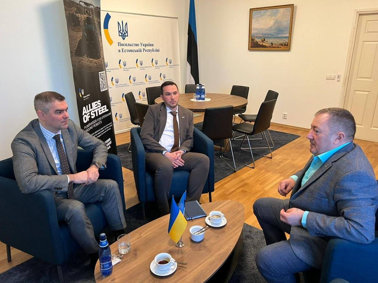 Троє чоловіків у костюмах ведуть переговори, сидячи в кріслах навколо журнального столика з напоями. Зустріч проходить у кабінеті на фоні банера Посольства України в Естонській Республіці та прапора Естонії. На стіні висить картина, а поруч стоїть круглий стіл для нарад.