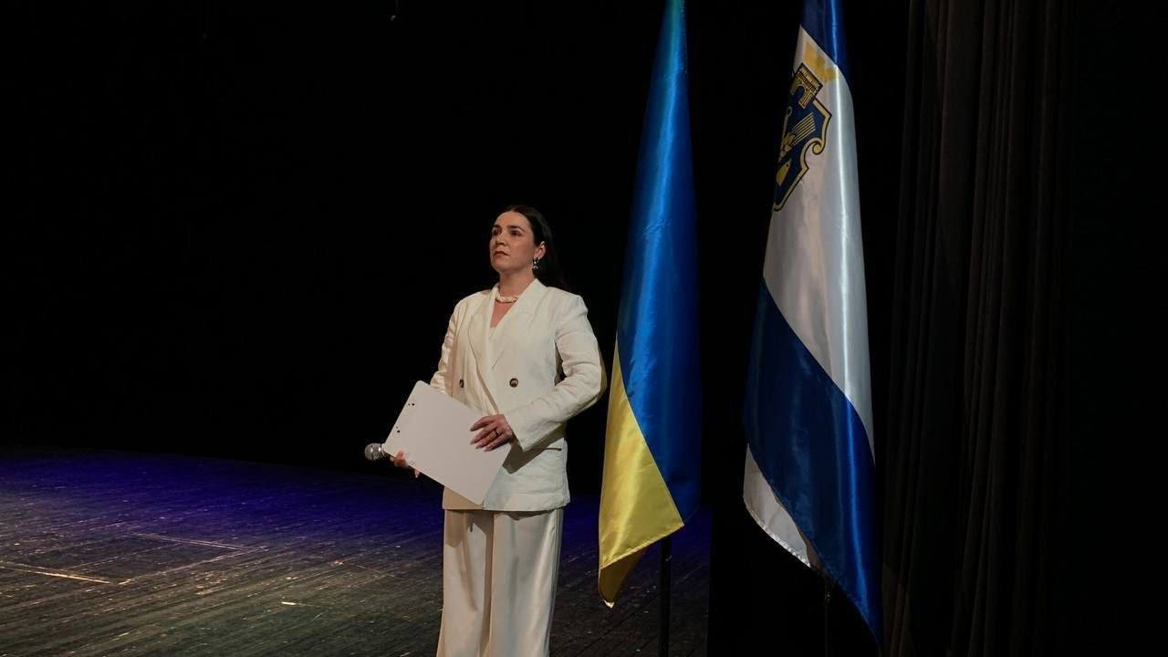 Жінка в білому костюмі стоїть на темній сцені, тримаючи в руках папку-планшет. Поруч із нею розташовані два прапори на флагштоках: державний прапор України та біло-синій прапор із гербом.