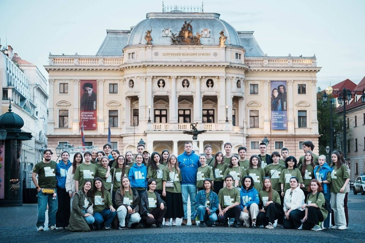Велика група молоді позує для спільного фото на площі перед величною історичною будівлею театру з куполом та колонами. Більшість учасників одягнені в однакові зелені та сині худі з написами. На фасаді будівлі розміщені великі театральні афіші.
