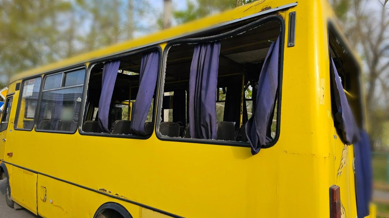 Жовтий пасажирський автобус із розбитим боковим та заднім склом. На вікнах видно пошкоджені сині шторки, а на корпусі помітні сліди руйнувань.
