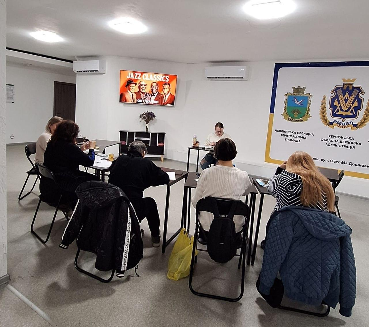 Група людей сидить за столами у світлому приміщенні під час робочої зустрічі або навчання. На стіні розміщено банер із гербами та телевізор, що транслює заставку «Jazz Classics».