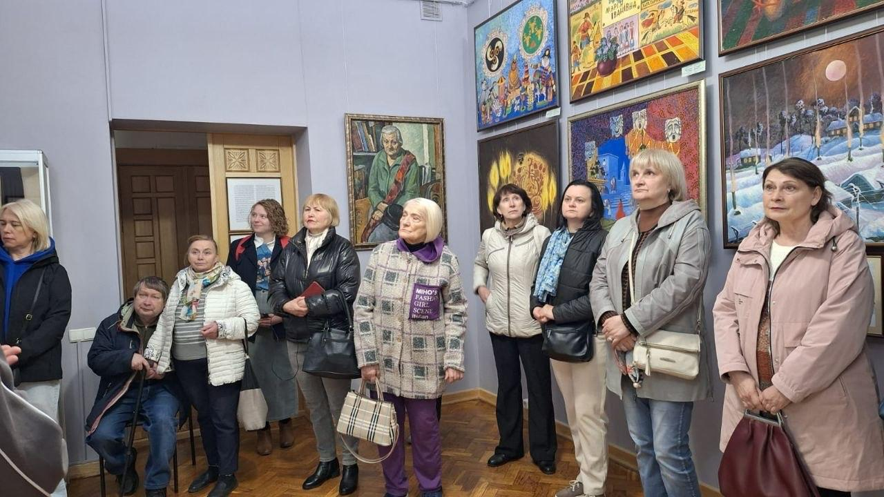 Група людей у верхньому одязі розглядає експозицію картин у художній галереї чи музеї. На стінах представлені різнопланові полотна — від портретів до сюжетних картин у яскравих рамах.