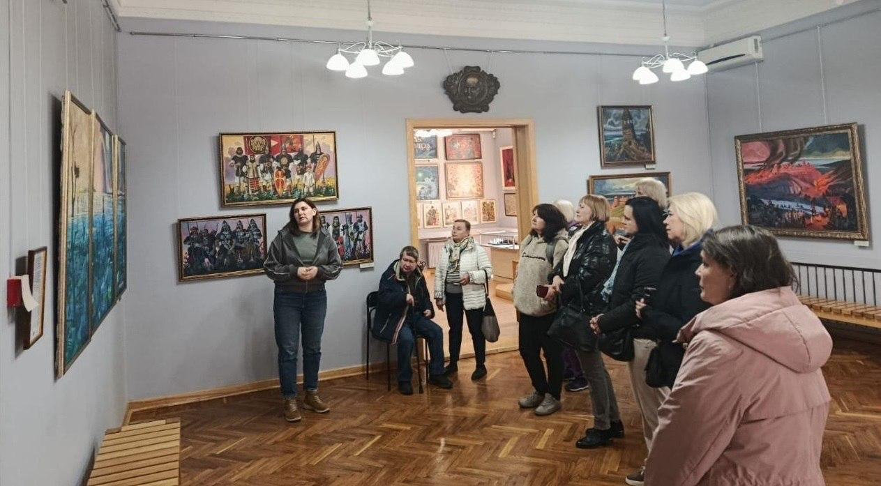 Група відвідувачів слухає екскурсовода у музейній залі. На стінах розміщені різноформатні картини, а крізь дверний отвір видно іншу залу з експозицією.