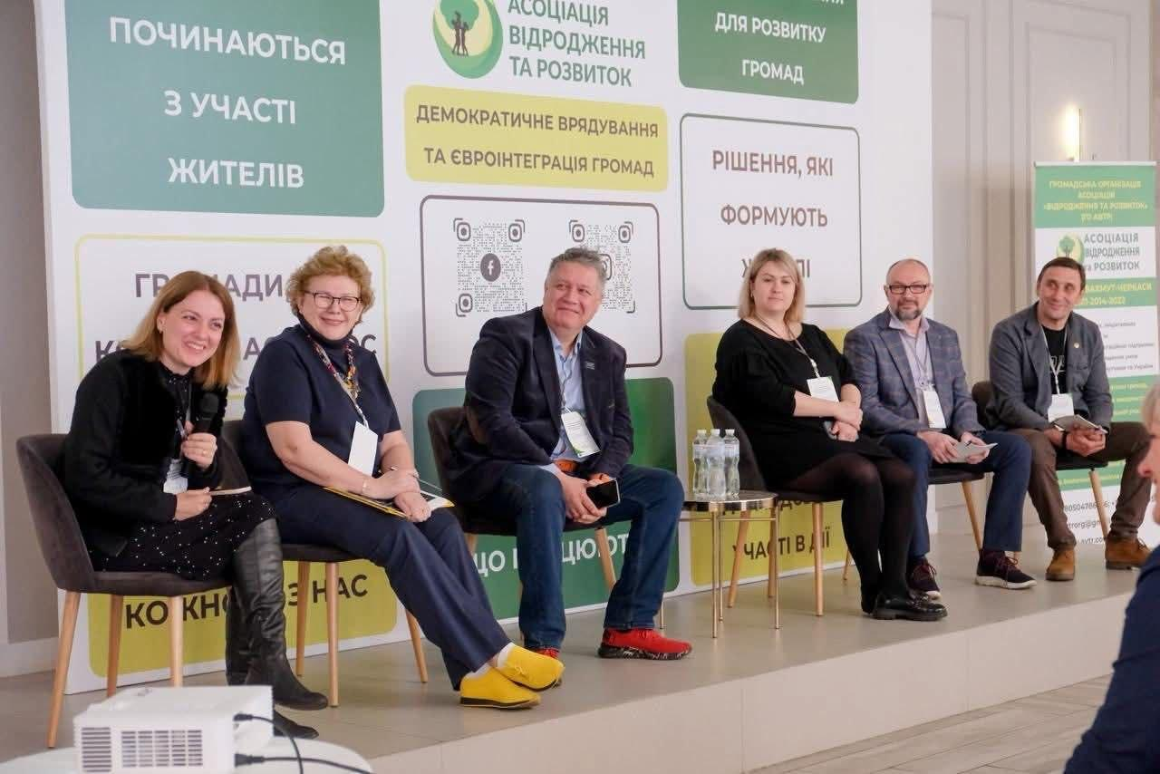 Шестеро спікерів сидять на сцені під час панельної дискусії на фоні великого банера з логотипом «Асоціація відродження та розвиток» та написами про демократичне врядування. Учасники посміхаються та тримають мікрофони, готуючись до виступу.