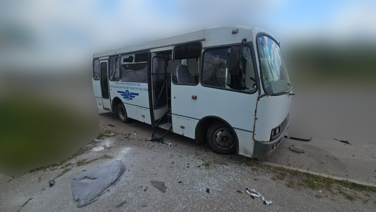Білий автобус із розбитим склом і відчиненими дверима стоїть на дорозі, навколо якої розкидані уламки.