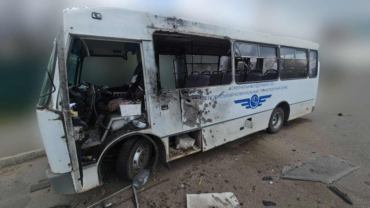 Білий комунальний автобус із численними отворами від уламків на корпусі та значними пошкодженнями передньої частини. У транспортного засобу вибито скло, а навколо на дорозі розкидані уламки.