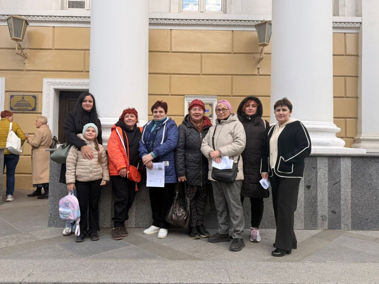 Група жінок та дівчинка стоять поруч для спільного фото на фоні будівлі з масивними білими колонами та жовтими стінами. Люди одягнені у демісезонний верхній одяг, дехто тримає в руках документи.