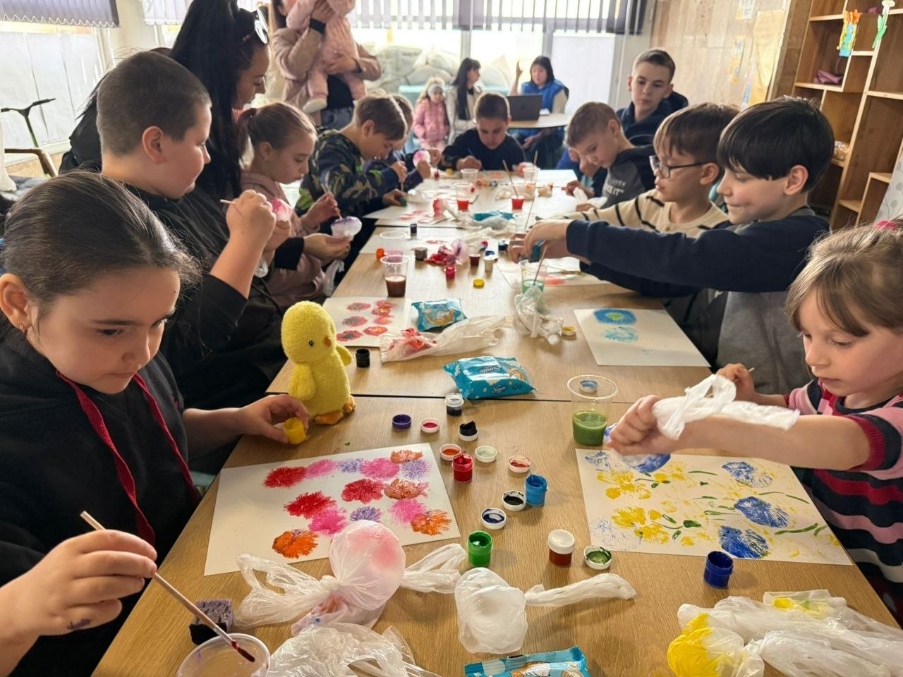Група дітей та дорослих сидить за довгим столом у світлій кімнаті, займаючись малюванням. Вони використовують фарби в баночках, пензлики та нетрадиційні матеріали, як-от зім’яті пакети, для створення яскравих малюнків на папері.