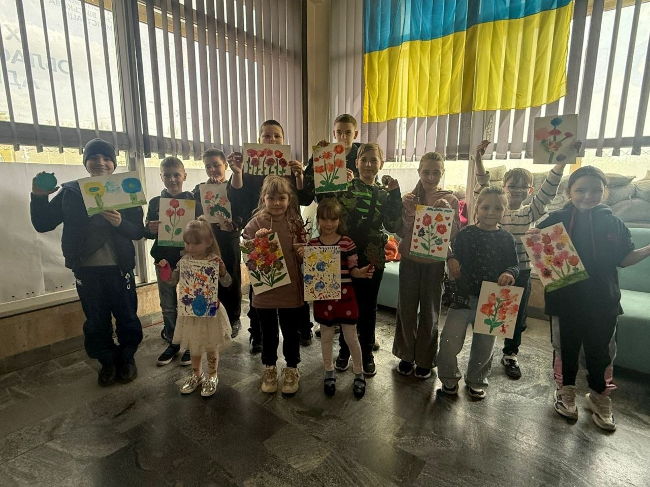 Група дітей різного віку стоїть у приміщенні та тримає перед собою свої малюнки з яскравими квітами. На задньому плані видно велике вікно з жалюзі та державний прапор України.