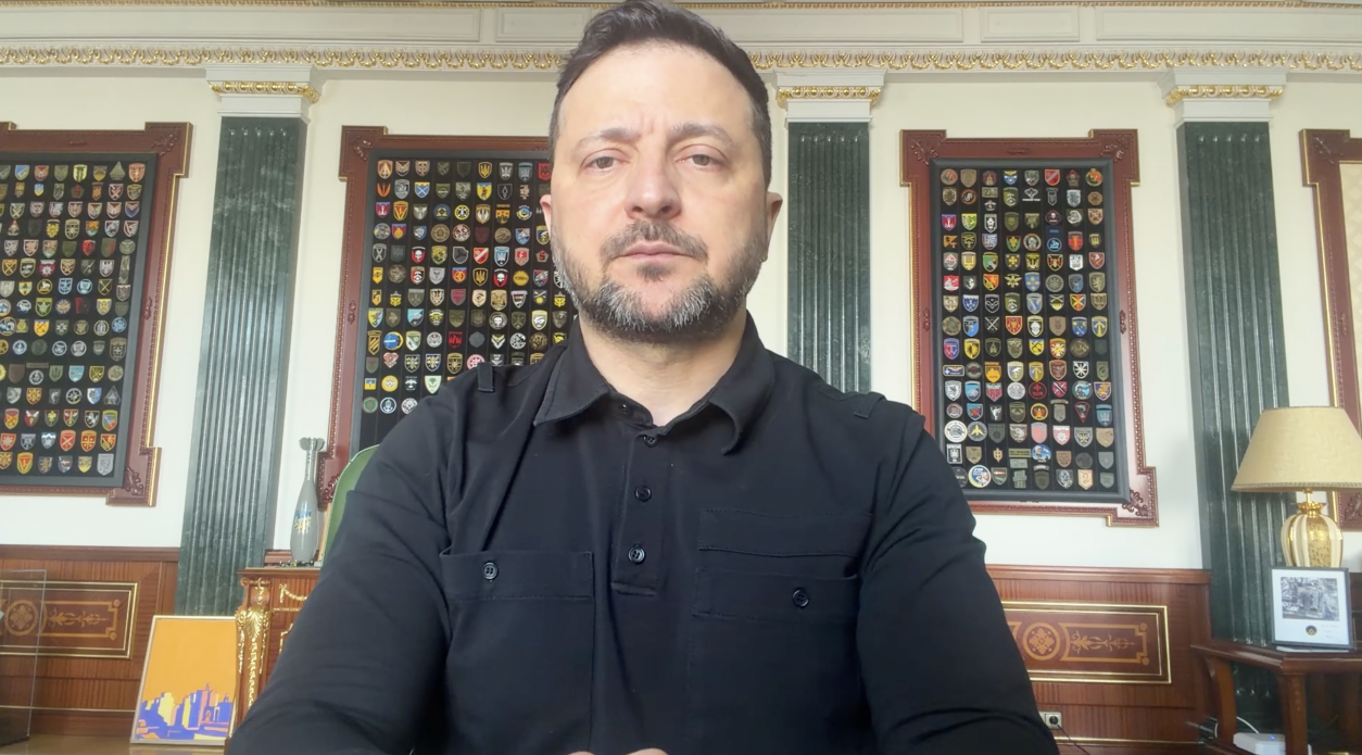 Чоловік у чорній сорочці сидить у кабінеті на фоні стін, декорованих колонами та стендами з численними військовими шевронами. Позаду нього також видно настільну лампу та елементи класичного інтер'єру.