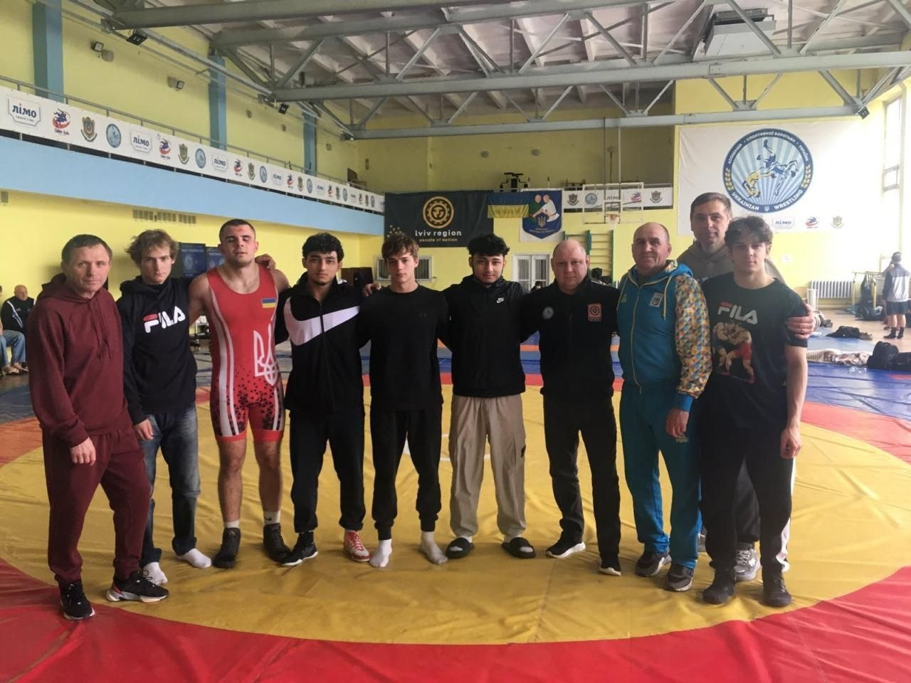 Група спортсменів та тренерів стоїть на борцівському килимі у спортивному залі. Один із юнаків одягнений у борцівське трико з гербом України, інші — у спортивні костюми та худі. На стіні залу видно банери та емблему спортивної федерації.