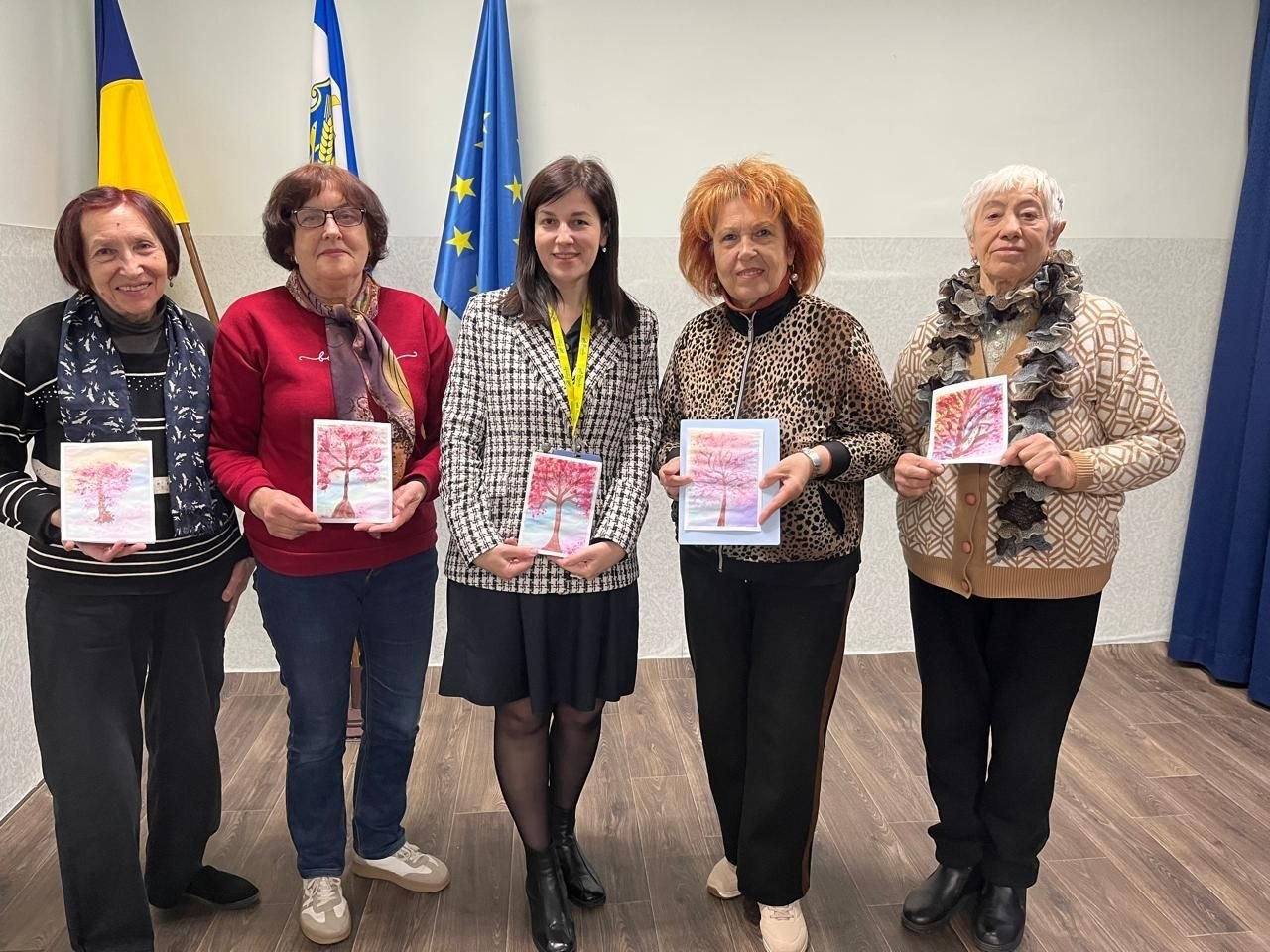 П'ять жінок стоять у ряд у приміщенні та тримають перед собою невеликі малюнки із зображенням квітучого дерева. На задньому плані стоять три прапори: державний прапор України, прапор міста та прапор Європейського Союзу.