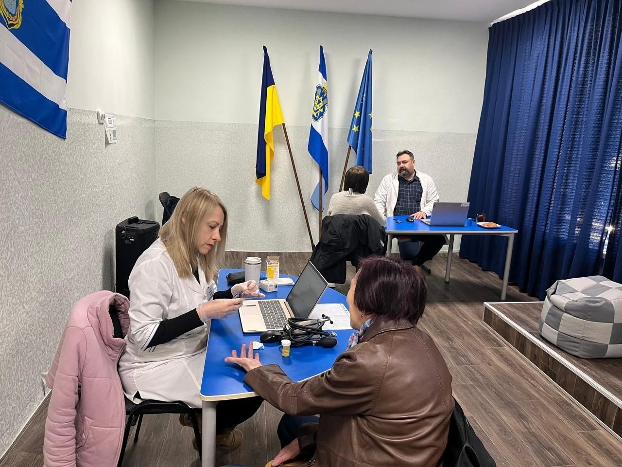 Медичні працівники у білих халатах приймають пацієнтів у кабінеті. На передньому плані медсестра проводить маніпуляцію на руці жінки, на задньому — лікар працює за ноутбуком. У приміщенні встановлені прапори України, міста та Європейського Союзу.