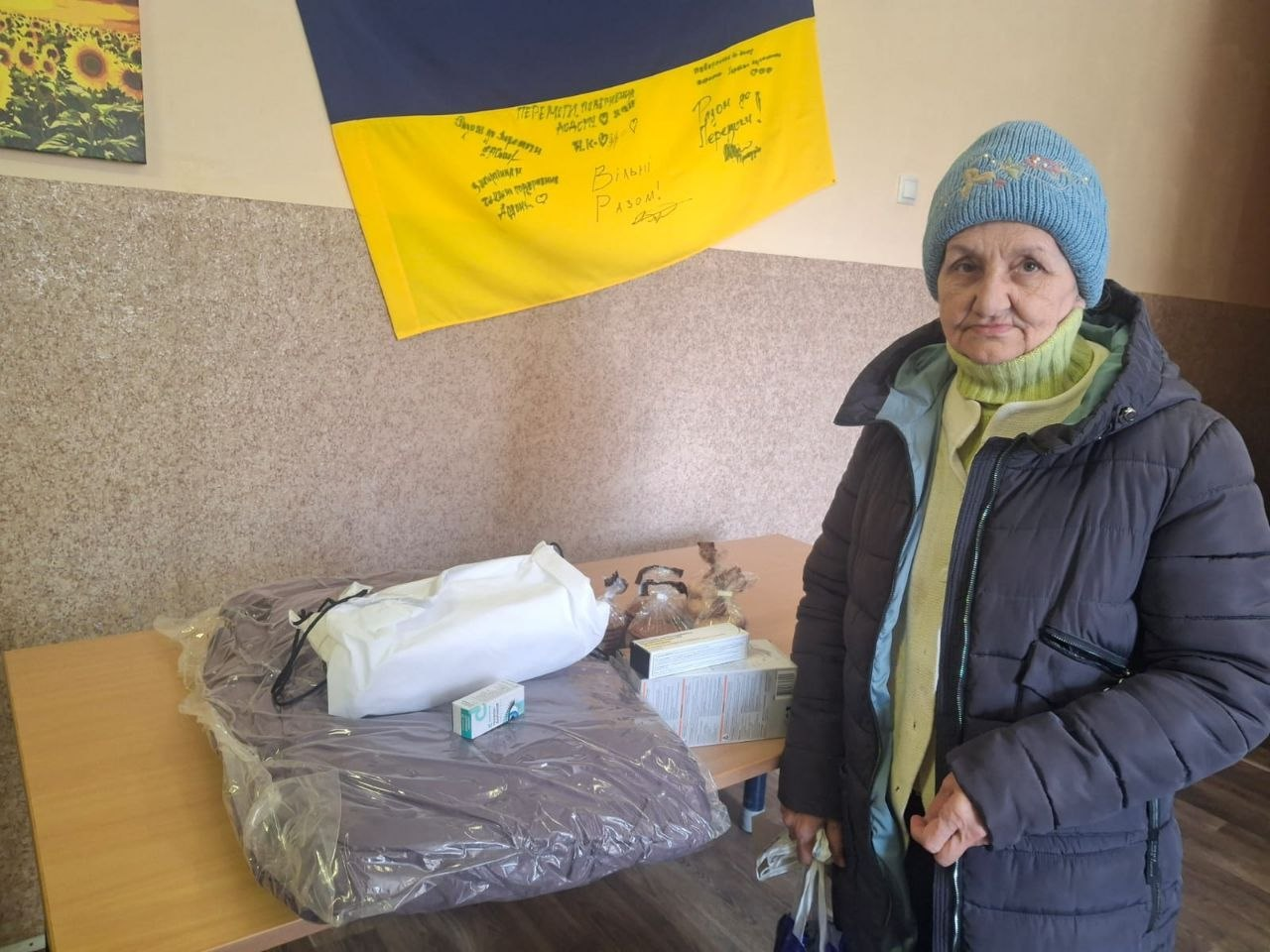 Літня жінка у теплому одязі стоїть поруч зі столом, на якому лежать пакунки з гуманітарною допомогою та ліками. На стіні за нею висить державний прапор України з написаними на ньому побажаннями.