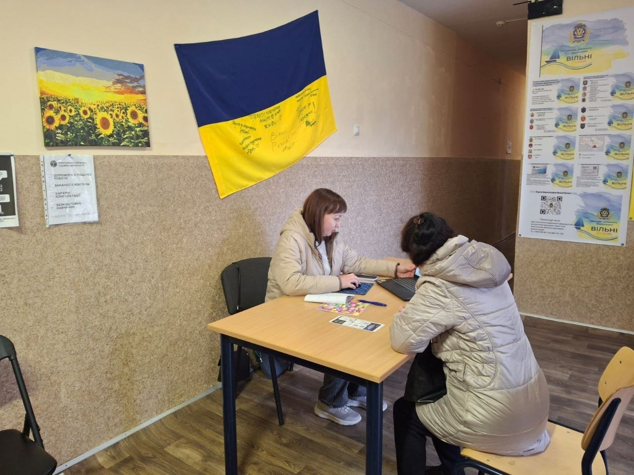 Жінка за робочим столом консультує відвідувачку, використовуючи ноутбук. На стіні за ними висить державний прапор України з написами, картина з соняшниками та інформаційні плакати.