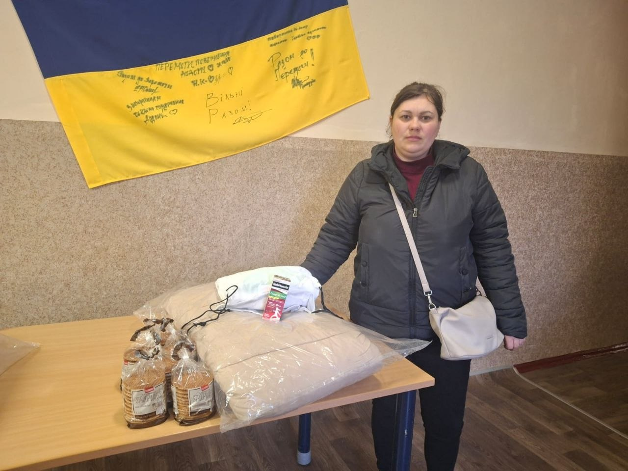 Жінка у темній куртці стоїть біля столу, на якому лежить пакунок з ковдрою та кілька пачок печива. На стіні за нею закріплений державний прапор України з рукописними написами та побажаннями.