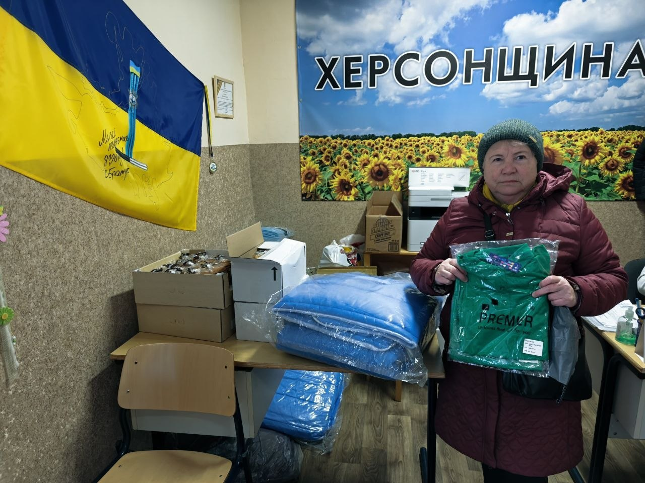 Жінка в бордовій куртці тримає пакунок із зеленим одягом. Поруч на столі лежать інші речі в упаковках та коробки. На стіні за нею висить банер «ХЕРСОНЩИНА» з полем соняшників та державний прапор України з малюнком і написами.