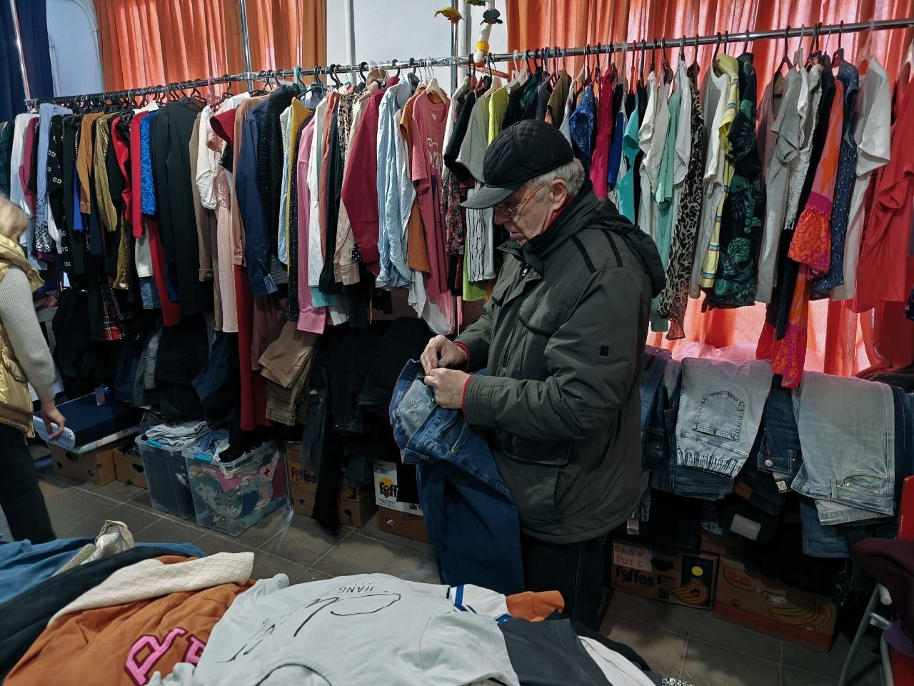 Чоловік у куртці та кепці розглядає сині джинси в приміщенні з великою кількістю вживаного одягу. Речі розвішані на довгих стійках та розкладені в коробках під ними.