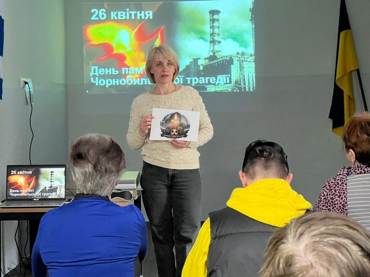 Жінка виступає перед аудиторією на тлі екрана з написом «26 квітня. День пам'яті Чорнобильської трагедії». Вона тримає в руках аркуш із тематичним малюнком, а перед нею сидять слухачі.