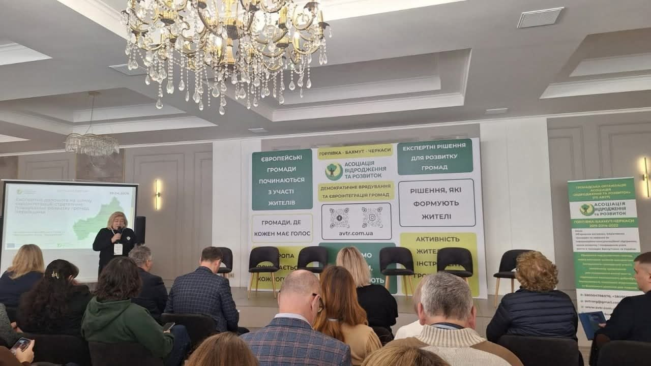 Жінка виступає з промовою перед аудиторією в офіційному залі. На задньому плані розміщено великий стенд «Асоціація відродження та розвиток» із текстом про європейські громади, а над залом звисає масивна кришталева люстра.