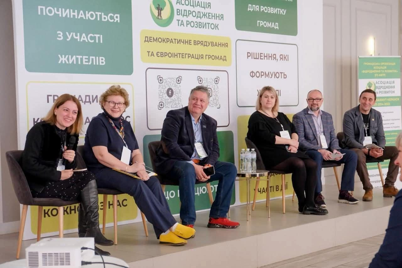 Шестеро спікерів сидять на сцені під час панельної дискусії на фоні банера з написами про демократичне врядування та розвиток громад. Учасники посміхаються, а на передньому плані видно частину мультимедійного проєктора.