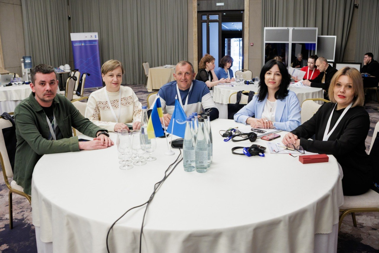 За круглим столом сидять п’ятеро учасників заходу: чоловік у зеленій сорочці, жінка у світлій мереживній блузі, чоловік у синьо-білому светрі, жінка у блакитному піджаку та жінка у чорному вбранні. Усі вони дивляться в кадр; на передньому плані перед ними стоять прапорці України та ООН.