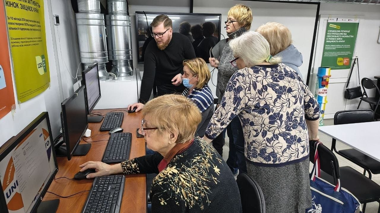 Група людей у комп'ютерному класі під час навчання. Кілька жінок старшого віку сидять за моніторами, а чоловік та ще одна жінка стоять поруч і допомагають їм опанувати техніку. На стінах розклеєні інформаційні плакати.