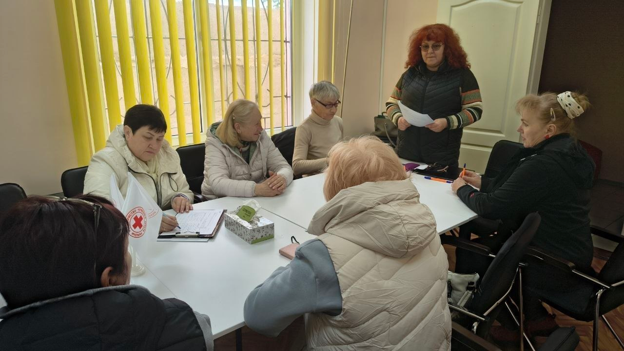 Група жінок проводить робочу зустріч у світлому кабінеті. П'ятеро учасниць сидять навколо столу, а одна стоїть і зачитує інформацію з аркуша. На столі стоїть маленький прапорець із логотипом Червоного Хреста, лежать папери для записів та серветки.