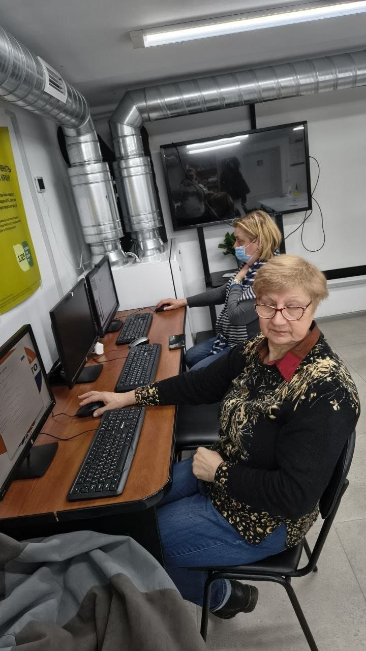 Дві жінки старшого віку працюють за комп’ютерами в навчальному класі. Вони зосереджено дивляться на монітори, тримаючи в руках комп’ютерні миші. На задньому плані видно вимкнений телевізійний екран та частину інформаційного плаката на стіні.