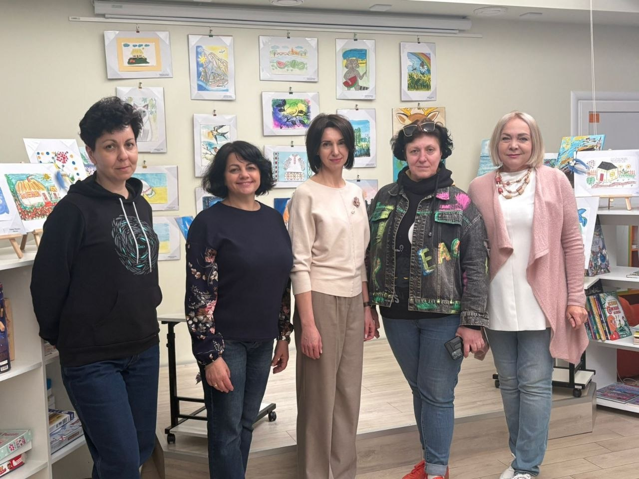 П'ятеро жінок стоять у ряд на фоні стіни, прикрашеної численними дитячими малюнками. Вони одягнені в кежуал-стилі: светри, джинси, джинсову куртку та кардиган. По боках від них видно стелажі з книгами та іграшками, що створює атмосферу освітнього або культурного центру.