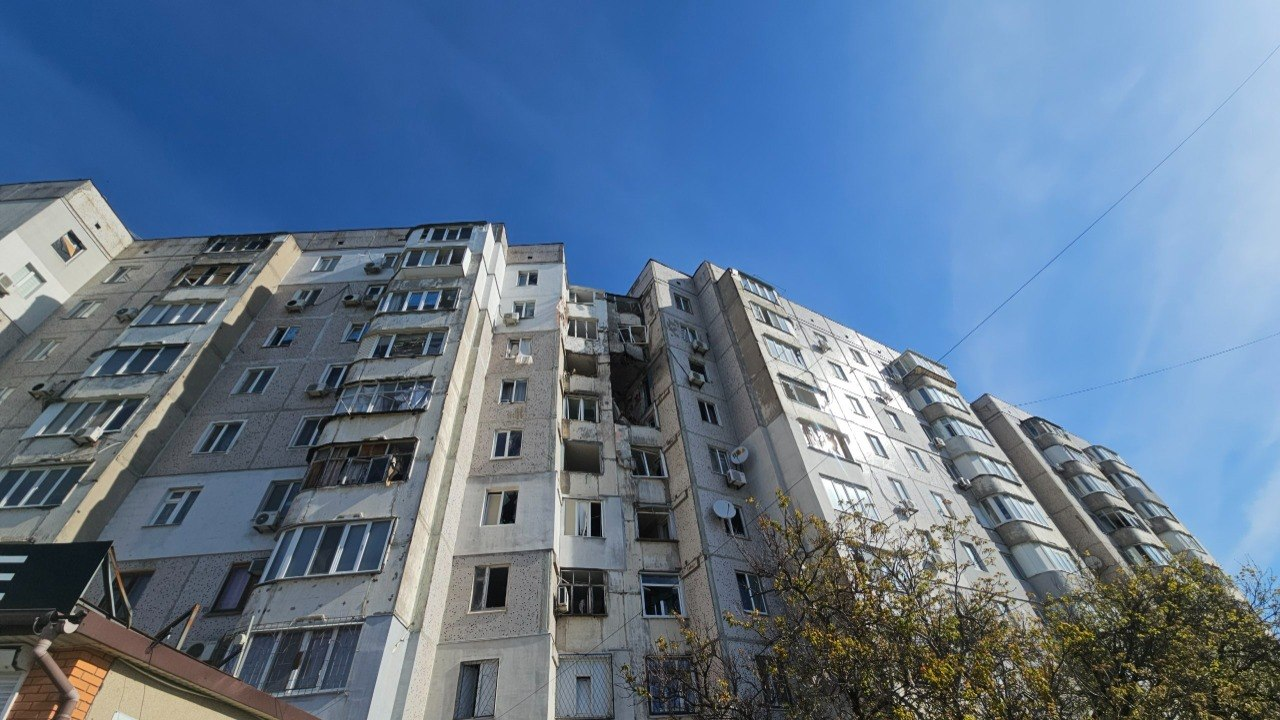 Пошкоджена багатоповерхівка з великою дірою у фасаді від влучання снаряда на тлі безхмарного синього неба.