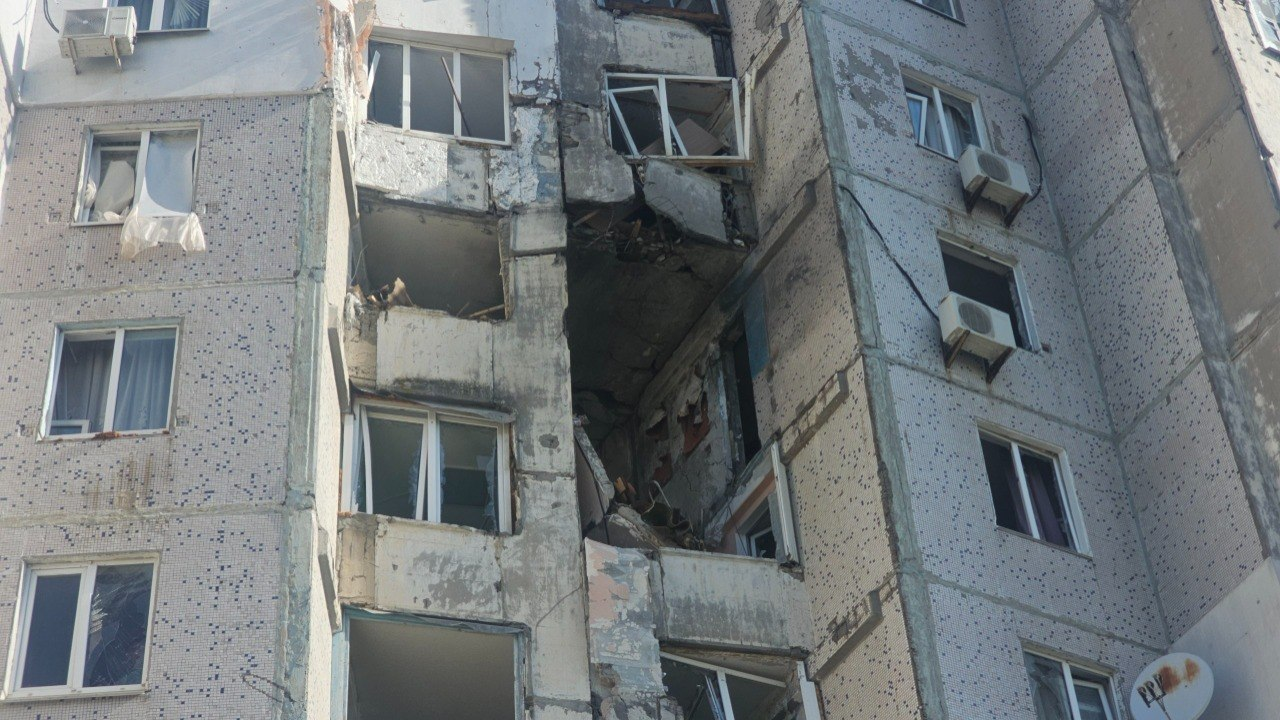 Фасад панельного будинку з наскрізною дірою від вибуху, вибитими вікнами та численними слідами від уламків на стінах.