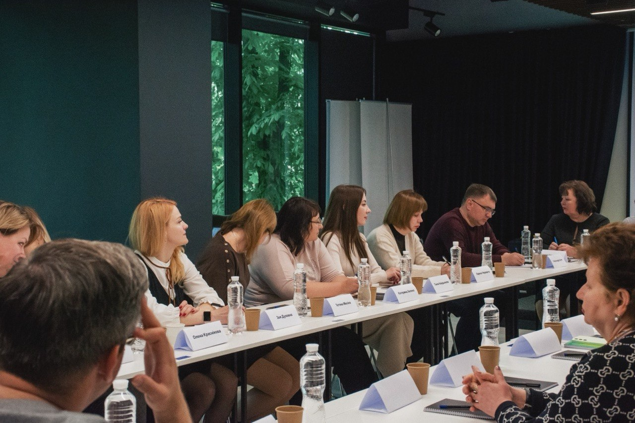 Група людей сидить за довгим білим столом під час ділової зустрічі або семінару в сучасному приміщенні з великими вікнами.