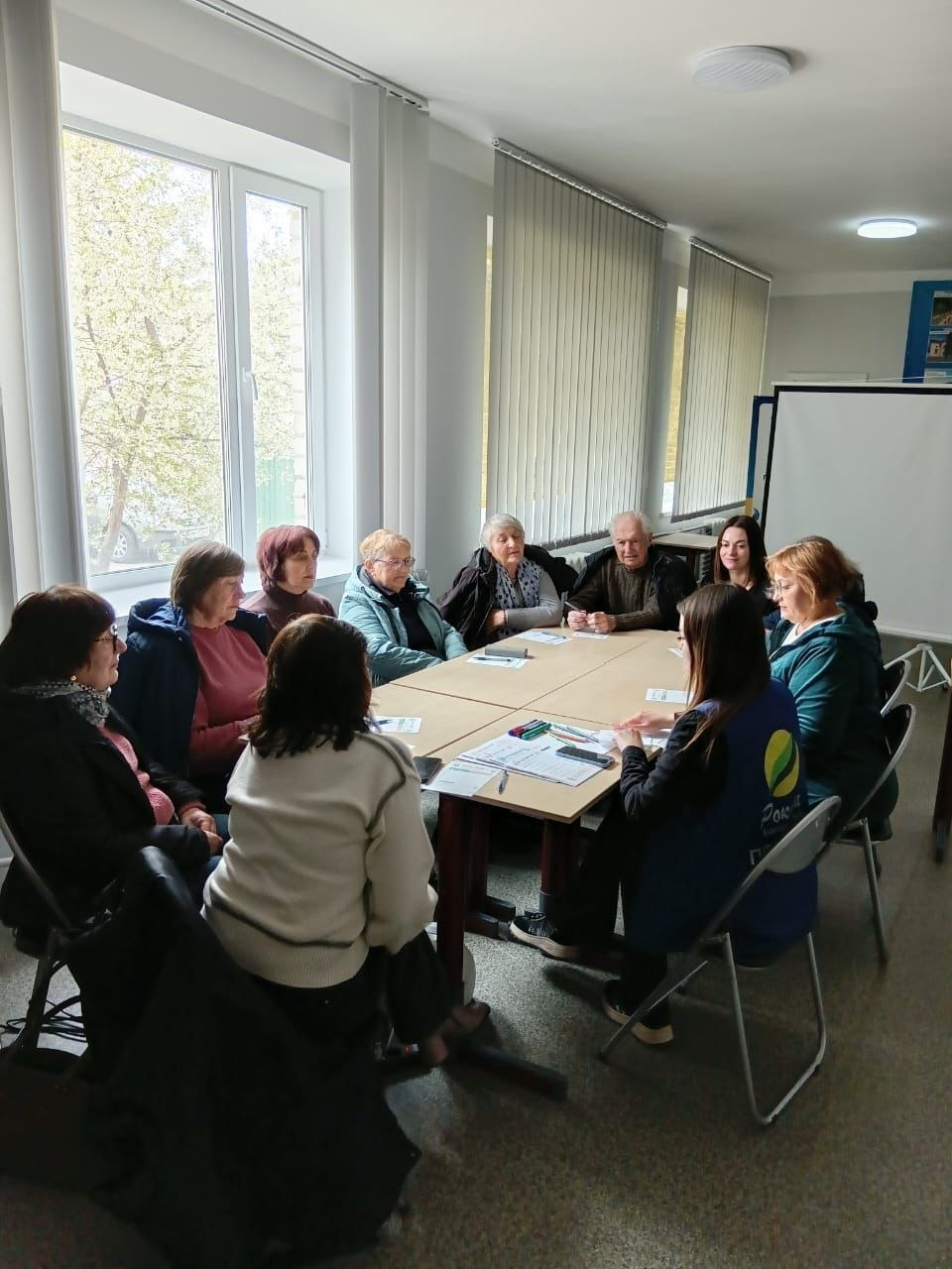 Група людей переважно похилого віку сидить за столом у світлій кімнаті під час зустрічі з представницею організації у синьому жилеті.