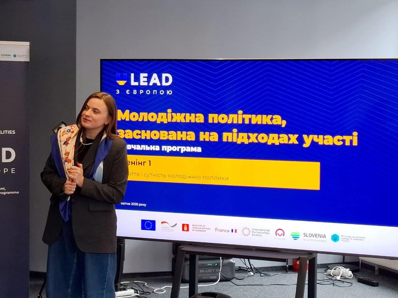 Жінка з букетом квітів стоїть поруч із великим екраном, на якому відображено заголовок презентації про молодіжну політику та логотип програми «LEAD з Європою».