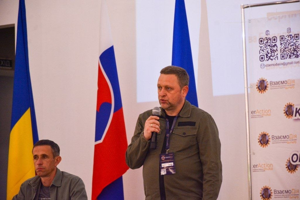 Чоловік у куртці кольору хакі виступає з мікрофоном на тлі державного прапора України та прапора з емблемою на білому фоні.
