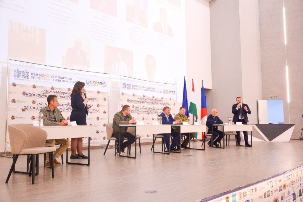 Жінка виступає з мікрофоном перед аудиторією на сцені IV Міжнародного конгресу «Взаємодія», де за столами також сидять інші учасники заходу.