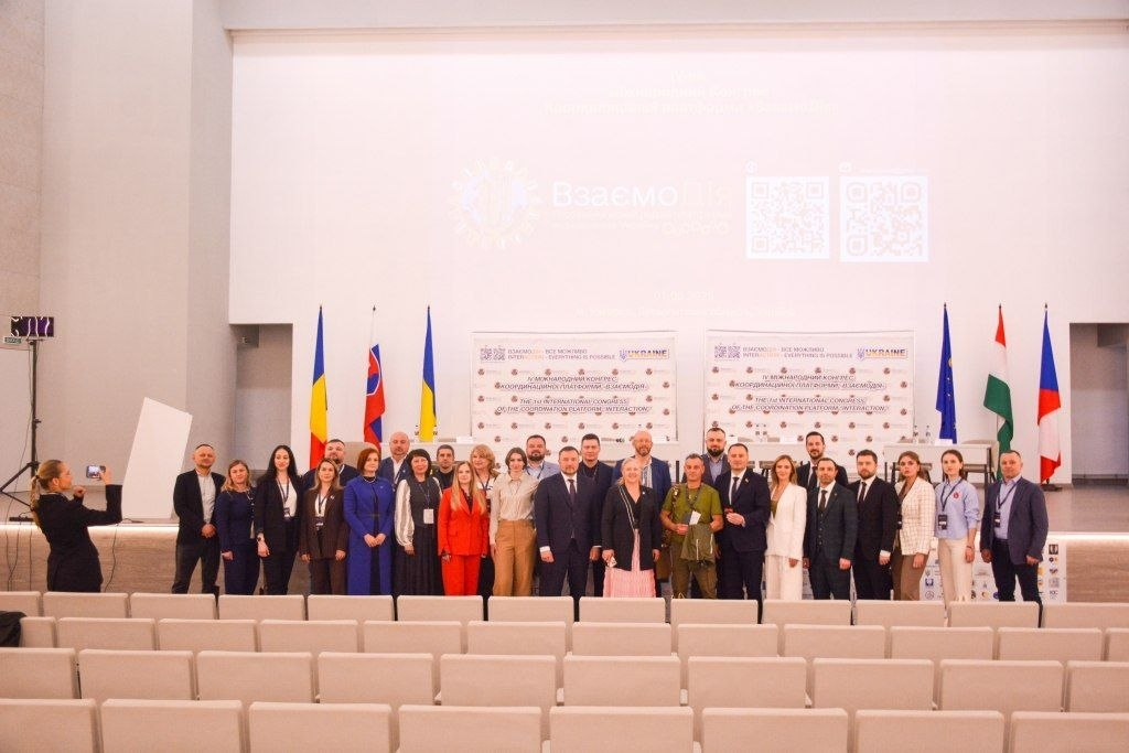 Велика група учасників позує для спільного фото на сцені IV Міжнародного конгресу «Взаємодія» на тлі банерів, державних прапорів та порожніх глядацьких рядів.