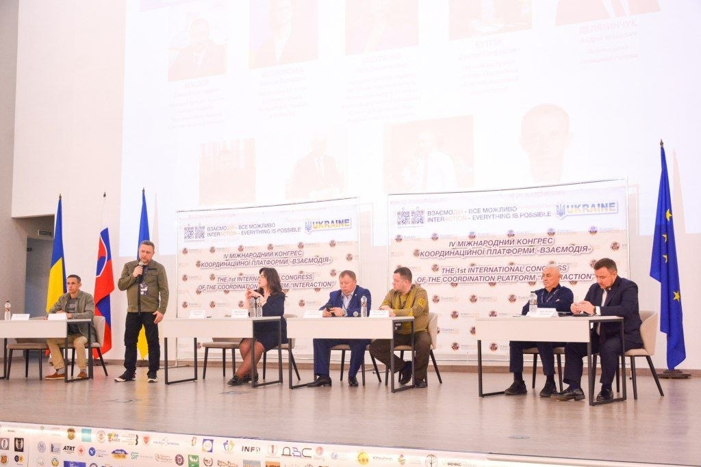 Чоловік у зеленому одязі виступає з мікрофоном на сцені офіційного заходу, де за столами сидять інші учасники на тлі банерів конгресу «Взаємодія».
