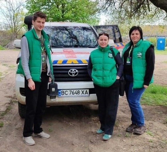 Троє людей у яскраво-зелених жилетах позують перед білим позашляховиком Toyota з українськими номерами на ґрунтовій дорозі поруч із деревами.