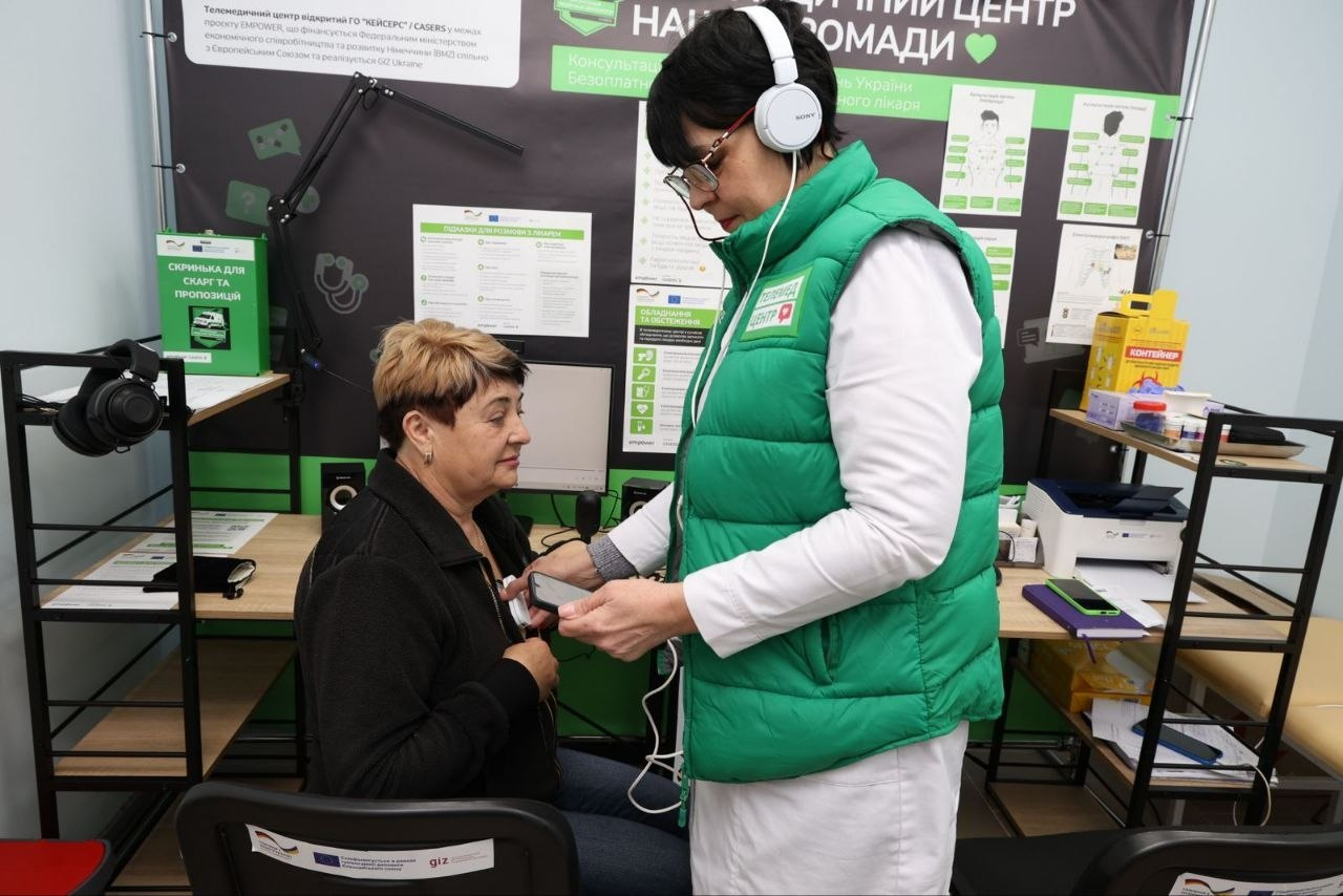 Медична працівниця в зеленому жилеті та навушниках проводить обстеження пацієнтки за допомогою цифрового пристрою в облаштованому кабінеті телемедицини.