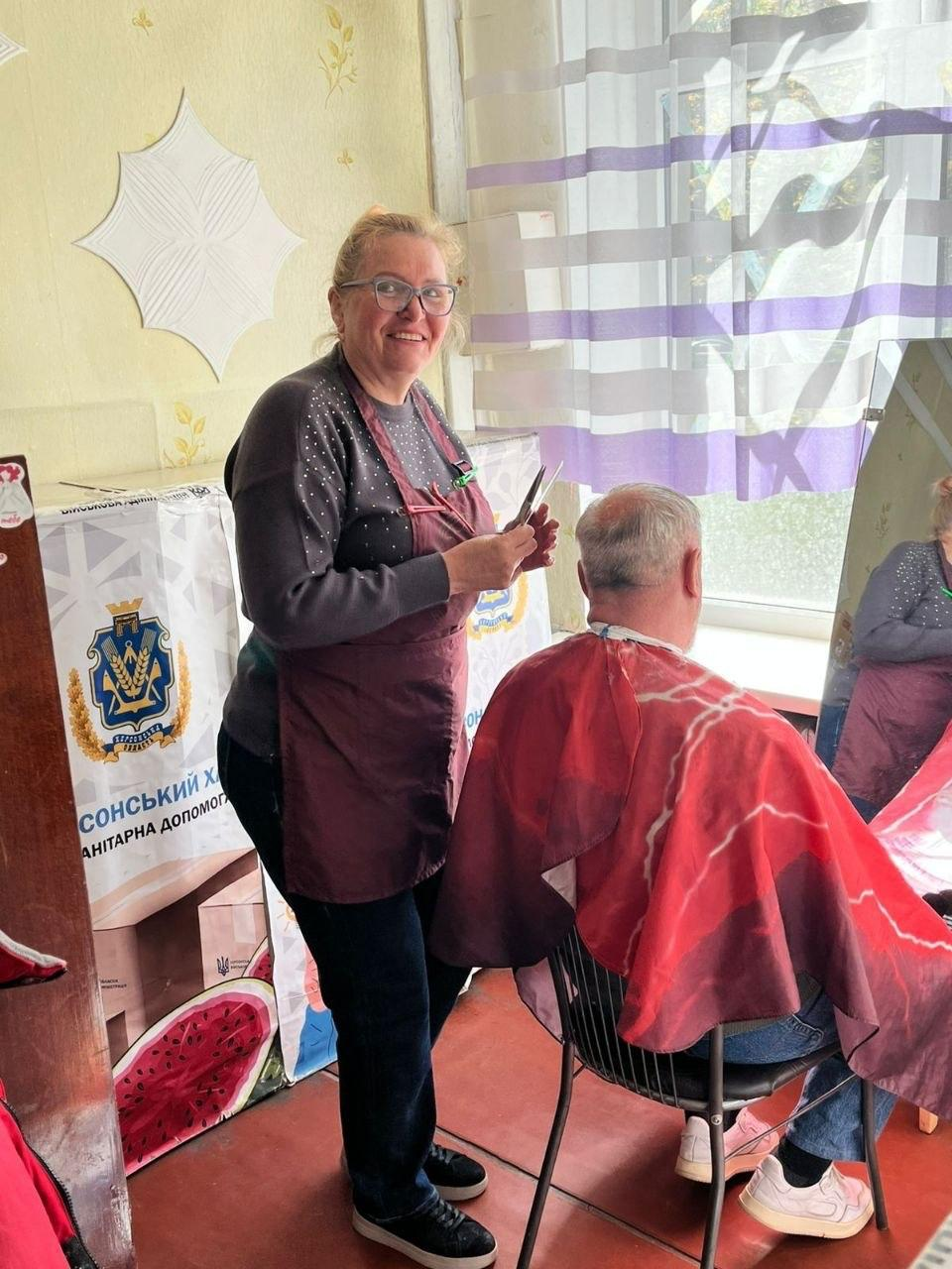 Жінка-перукар у бордовому фартуху стриже чоловіка в облаштованому приміщенні з логотипом Херсонського хабу гуманітарної допомоги на стіні.