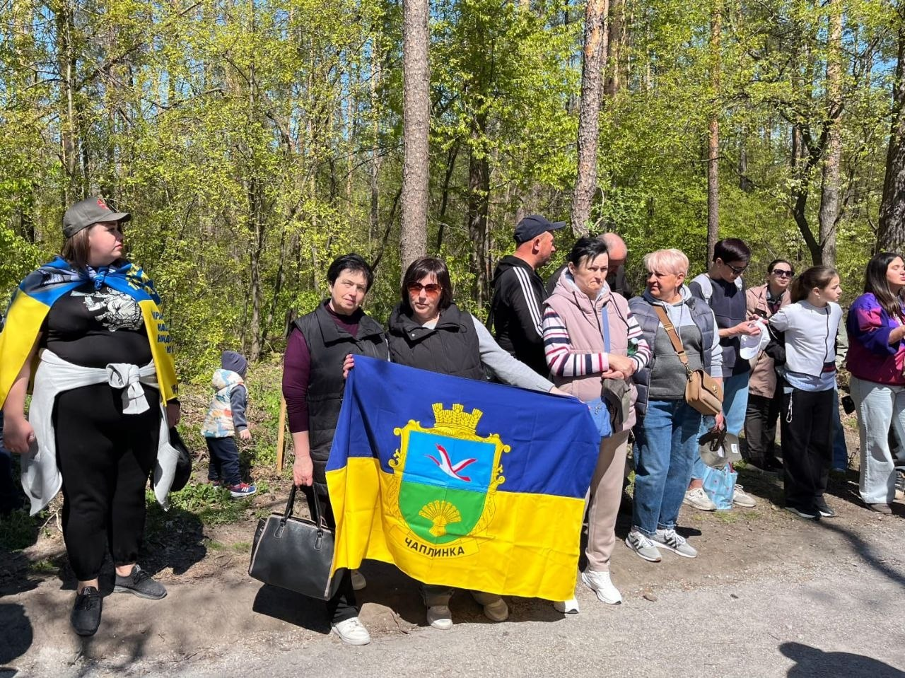 Група людей стоїть на узліссі; у центрі дві жінки тримають синьо-жовтий прапор із гербом та написом «Чаплинка» на тлі молодих весняних дерев.